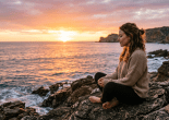 Woman sitting cross-legged on rocks by ocean at sunset